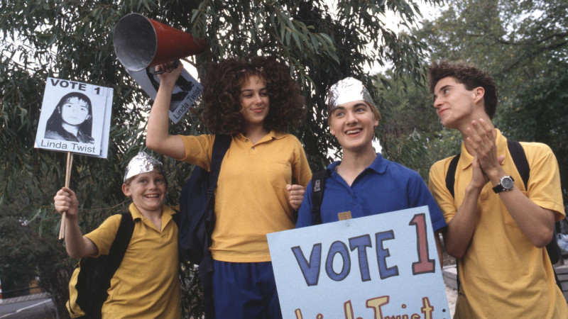 RTT_S4_EP07_2394 A group of four children stands outdoors, rallying for a campaign. One girl holds a megaphone high and wears a cheering expression, while another girl displays a sign with a photo of "Linda Twist." Two boys, one wearing a tinfoil hat, hold signs supporting the same candidate, smiling and looking enthusiastic. They are dressed in bright yellow and blue shirts.