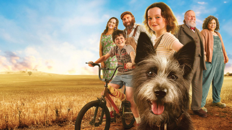 A group of six characters stands in a sunlit field, with a young girl and a dog in the foreground. The girl is smiling and riding a bicycle, while the others, including adults and children, display various expressions of joy and curiosity. The landscape features golden fields and a blue sky, enhancing the cheerful atmosphere.
