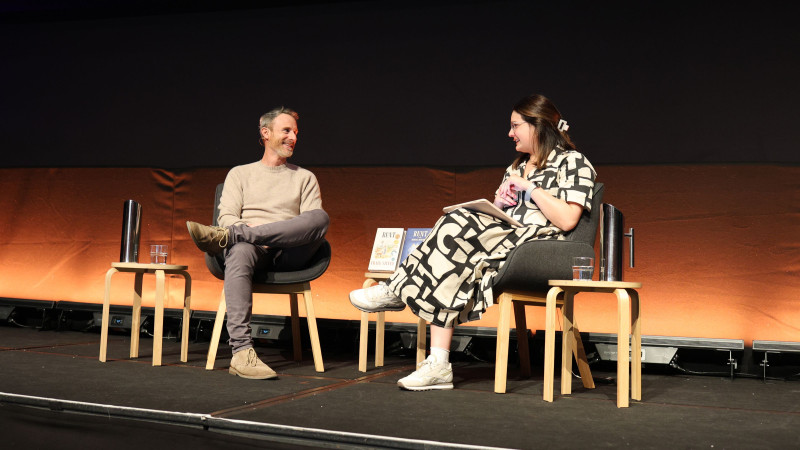 A man and a woman are seated on stage, engaged in conversation. The man, dressed in a light sweater and gray pants, smiles as he listens, while the woman, wearing a black and white patterned dress, holds a notebook. Two wooden stools hold a water glass and a metal pitcher, along with two books displayed on the between them. The background features an orange wall and a dimly lit ambiance.