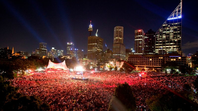 A large crowd gathers at a nighttime concert, with numerous people illuminated by red lights. In the background, a skyline of tall buildings and structures, including a prominent tower and various skyscrapers, is visible against the dark sky.