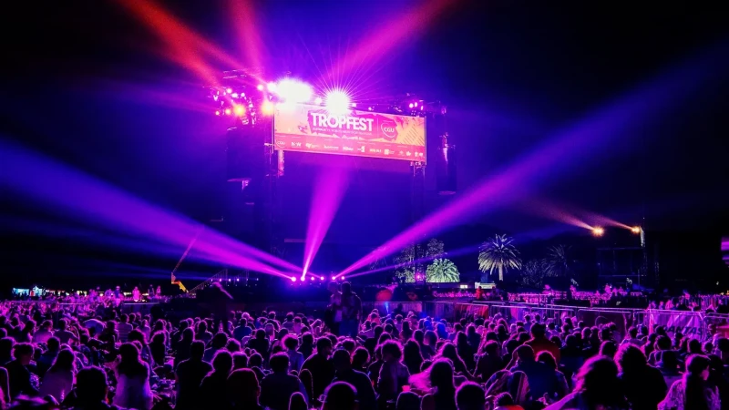 A large outdoor crowd gathers under colorful lights at a nighttime festival, with a prominent stage displaying a banner for Tropfest. The atmosphere is vibrant, enhanced by beams of purple and orange lights illuminating the scene, while palm trees can be seen in the background.
