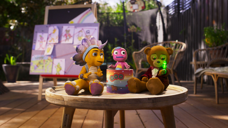 A wooden table in an outdoor setting features three colorful plush characters: a yellow creature with a floral headpiece, a pink figure in a dress, and a bear wearing a red jacket with a glowing green object. Behind them, drawings are pinned to a board, adding a playful ambiance to the scene.