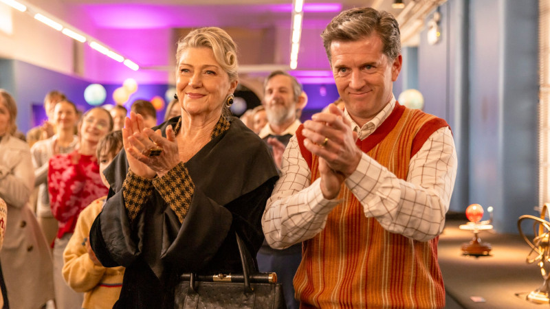 A woman with styled hair and elegant attire smiles while clapping her hands, standing beside a man in a colorful, retro sweater who also claps. They are joined by a crowd in a festive setting, with colorful lights illuminating the background. The atmosphere appears celebratory and joyful.