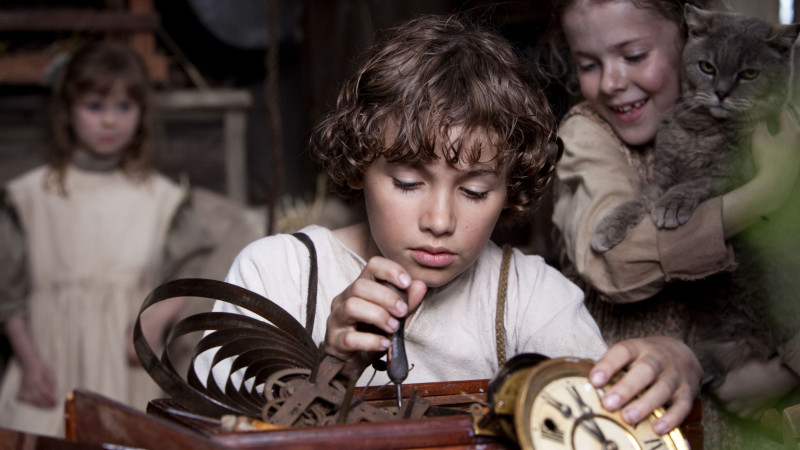 A young boy with curly hair examines a wooden clock mechanism, using a small tool with concentration. In the background, two girls, one holding a cat, watch him with smiles, creating a sense of camaraderie in the cozy, rustic setting.