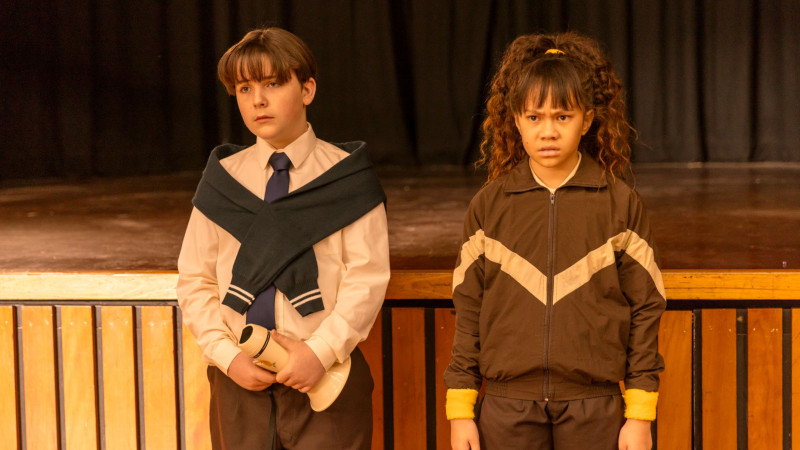 Two children stand side by side on a stage, looking serious. One boy wears a white shirt with a tie, a navy sweater draped over his shoulders, and holds a rolled-up paper, while the girl is dressed in a brown tracksuit, her expression tense, with her arms crossed in front of her. The background is dark with a wooden stage in front of them.