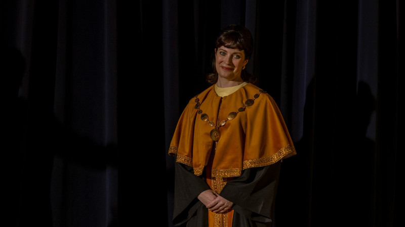 A woman stands confidently on stage, wearing a distinctive ceremonial outfit featuring an orange cape with golden embellishments. She has a pleasant smile and her hair is styled in loose curls, with the dark backdrop creating a dramatic contrast.