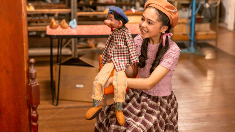 A young girl with long hair in pigtails wears a light pink top and a tan beret, smiling as she holds a puppet resembling a boy dressed in plaid and trousers. The background features wooden floors and shelves filled with various items, creating a playful and nostalgic atmosphere.