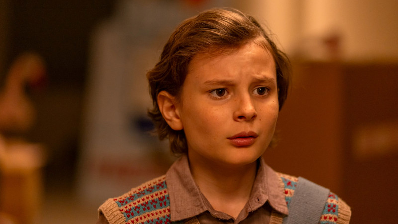 A young boy with short, wavy hair looks concerned, displaying a worried expression on his face. He wears a patterned vest over a collared shirt, set against a softly lit background with hints of indistinct shapes and colors.