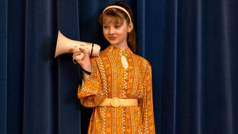 A girl wearing a bright, vintage-style dress with orange and yellow patterns stands confidently against a deep blue curtain, holding a megaphone in her right hand. She has a playful expression and her hair is styled with a headband, enhancing her retro look.