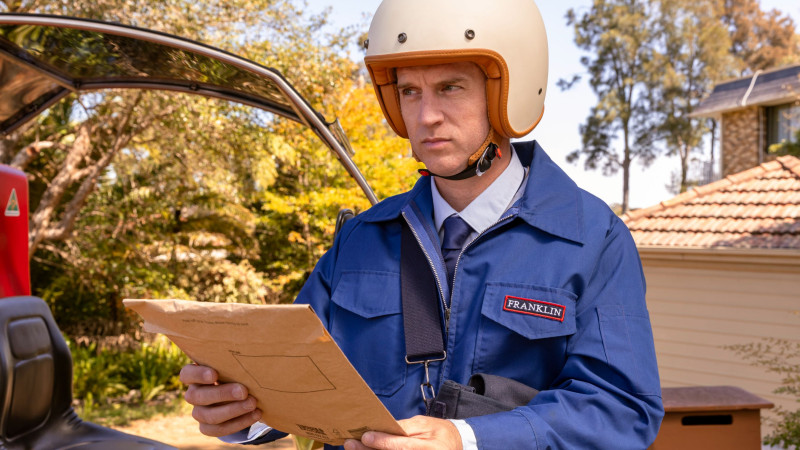 A man wearing a beige helmet and a blue uniform with a "FRANKLIN" patch examines a brown envelope in his hands. He stands next to a red vehicle, surrounded by greenery and a residential area in the background.