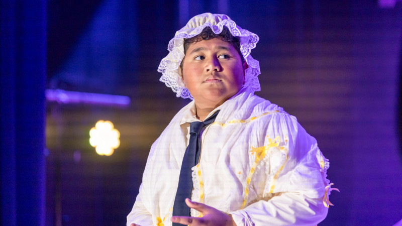 A young boy wearing a white dress and bonnet stands on stage, looking serious. He also sports a black tie, and the background features soft, colorful lighting that suggests a performance setting.