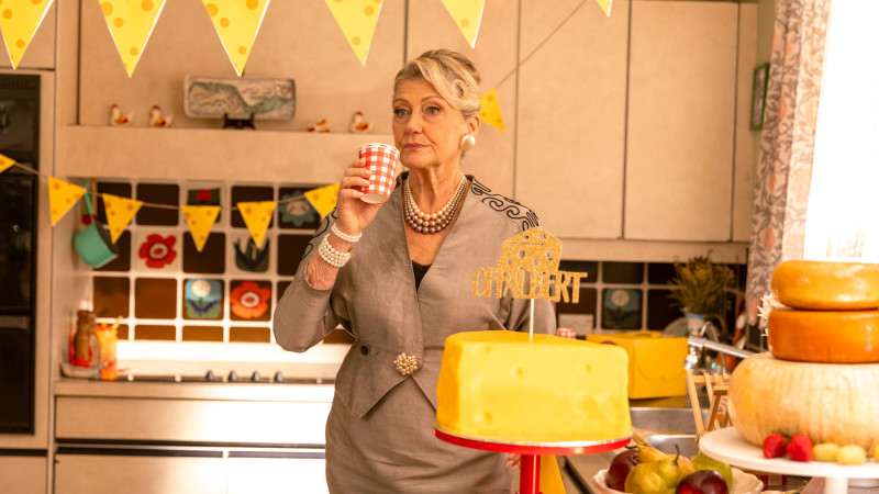 A woman in a stylish gray outfit with pearls stands in a brightly decorated kitchen, holding a red and white checkered cup. The kitchen features yellow cheese-themed decorations and a large yellow cake on a stand, accompanied by various types of cheese and fruits, all set against a backdrop of colorful bunting and floral tiles.