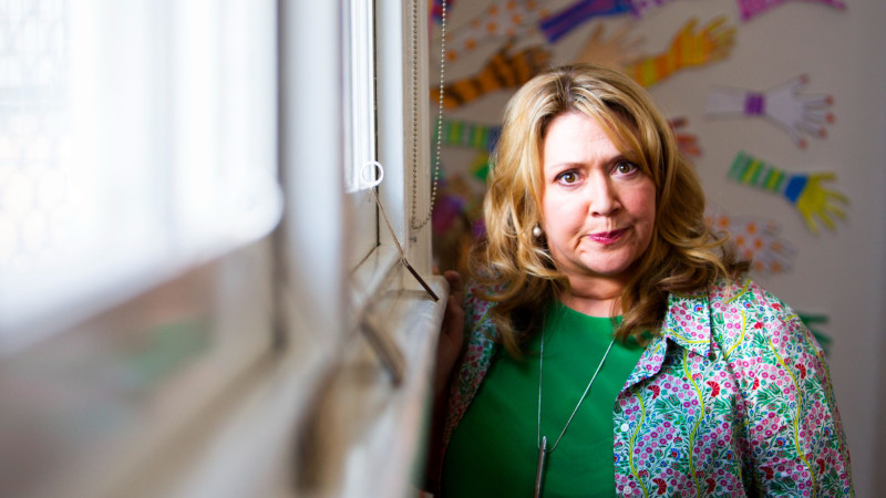 A woman with wavy blonde hair stands near a window, wearing a green shirt and a colorful floral-patterned cardigan. She looks directly at the camera with a serious expression, and a wall behind her features a playful display of colorful handprints.