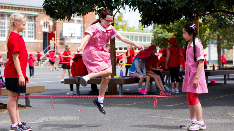 A girl in a pink dress and glasses jumps rope while another girl in a red shirt stands by, watching. In the background, other children in red clothing are engaged in various activities, and benches are visible under leafy trees.