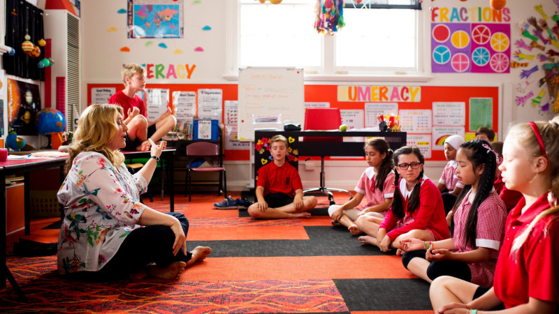 A group of children sits cross-legged on a colorful carpet in a classroom, engaged in meditation. A teacher, seated nearby and gesturing, guides them as a boy balances on a chair in the background. Brightly colored educational posters adorn the walls, and various classroom supplies are visible on tables.