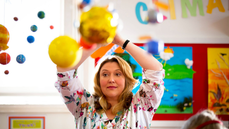 A woman with blonde, wavy hair is in a colorful classroom filled with hanging decorations of planets and celestial bodies. She is reaching upwards with a focused expression, while vibrant artwork adorns the walls behind her.