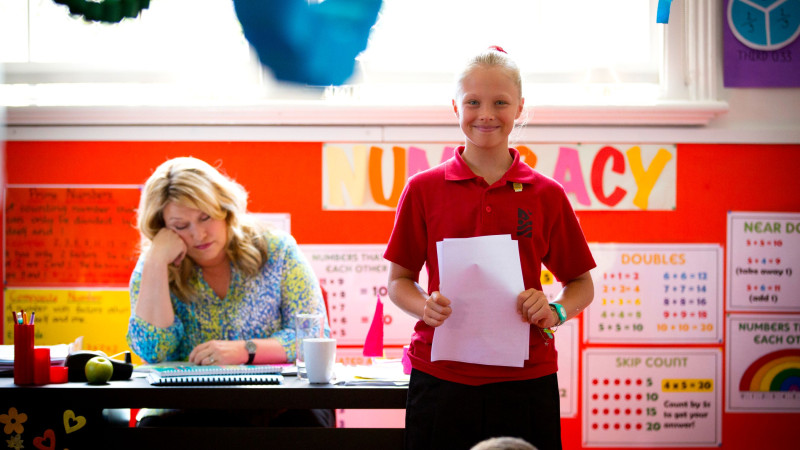 A young girl in a red polo shirt stands confidently in front of a classroom, holding a sheet of paper and smiling. In the background, a woman with blonde hair rests her head on her hand, seemingly overwhelmed, while colorful educational posters decorate the bright walls.