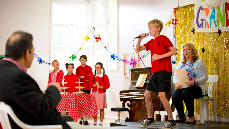 A young boy in a red shirt performs on stage with a microphone, while seated adults and a group of children in red outfits watch intently. The backdrop features colorful decorations and a banner, creating a festive atmosphere. The audience, including a man and a woman, display expressions of interest and surprise.