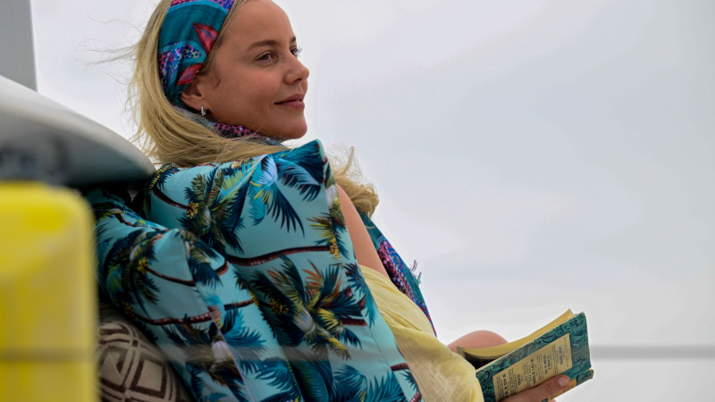 A woman with long, blonde hair, wearing a colorful headband and a tropical-patterned jacket, sits relaxed while holding a book. The background features a cloudy sky and the hint of a boat, creating a serene atmosphere.