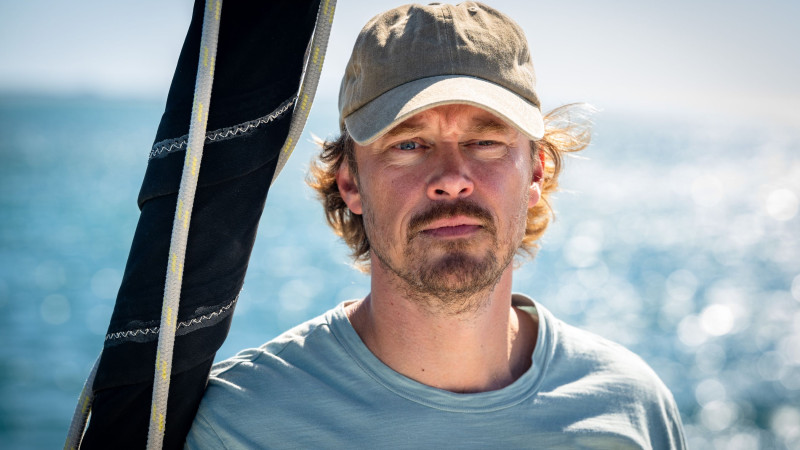 A man with tousled hair and a light blue shirt stands near the water, looking directly at the camera with a serious expression. He wears a beige cap and has a backdrop of shimmering blue waves.