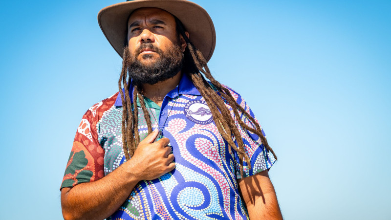A man with long dreadlocks stands against a clear blue sky, wearing a colorful shirt adorned with traditional patterns. He holds one hand over his heart, exuding a sense of pride and connection to his heritage.