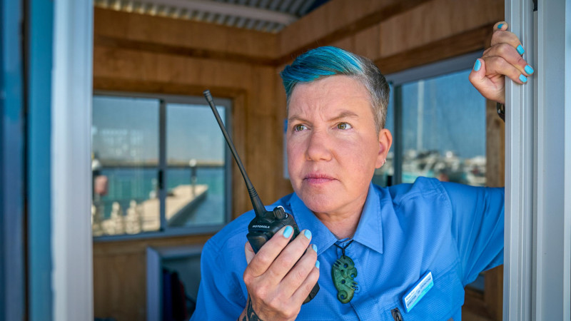 A person with short, dyed blue hair holds a two-way radio, looking intently as they stand in a wooden structure with large windows. They wear a blue uniform shirt and display visible tattoos on their arm, suggesting a focused and alert demeanor while communicating.