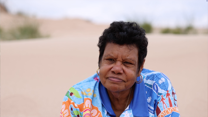 A woman with short, curly black hair sits in a sandy environment, wearing a brightly colored shirt with floral and geometric patterns. She has a serious expression and is surrounded by a blurred background of sand and greenery.
