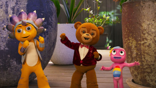 Three colorful characters are posed together in a bright outdoor setting. One character is orange with a large spiked head, another is a bear wearing a red tuxedo jacket, and the third is a pink figure with a rainbow design on its chest, all looking cheerful. Surrounding them are large planters and greenery, adding a vibrant backdrop.