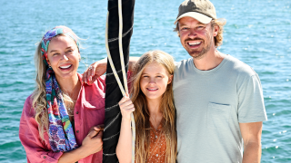 A woman with long light hair wearing a colorful scarf stands beside a girl who is holding onto a rope; both smile at the camera. To the right, a man with a cap and a light shirt grins, creating a cheerful family scene by the water.
