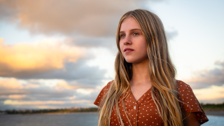 A girl with long, wavy blonde hair stands outdoors, gazing into the distance with a thoughtful expression. She wears a brown polka-dotted dress and is surrounded by a scenic backdrop of clouds and a soft-lit sky.