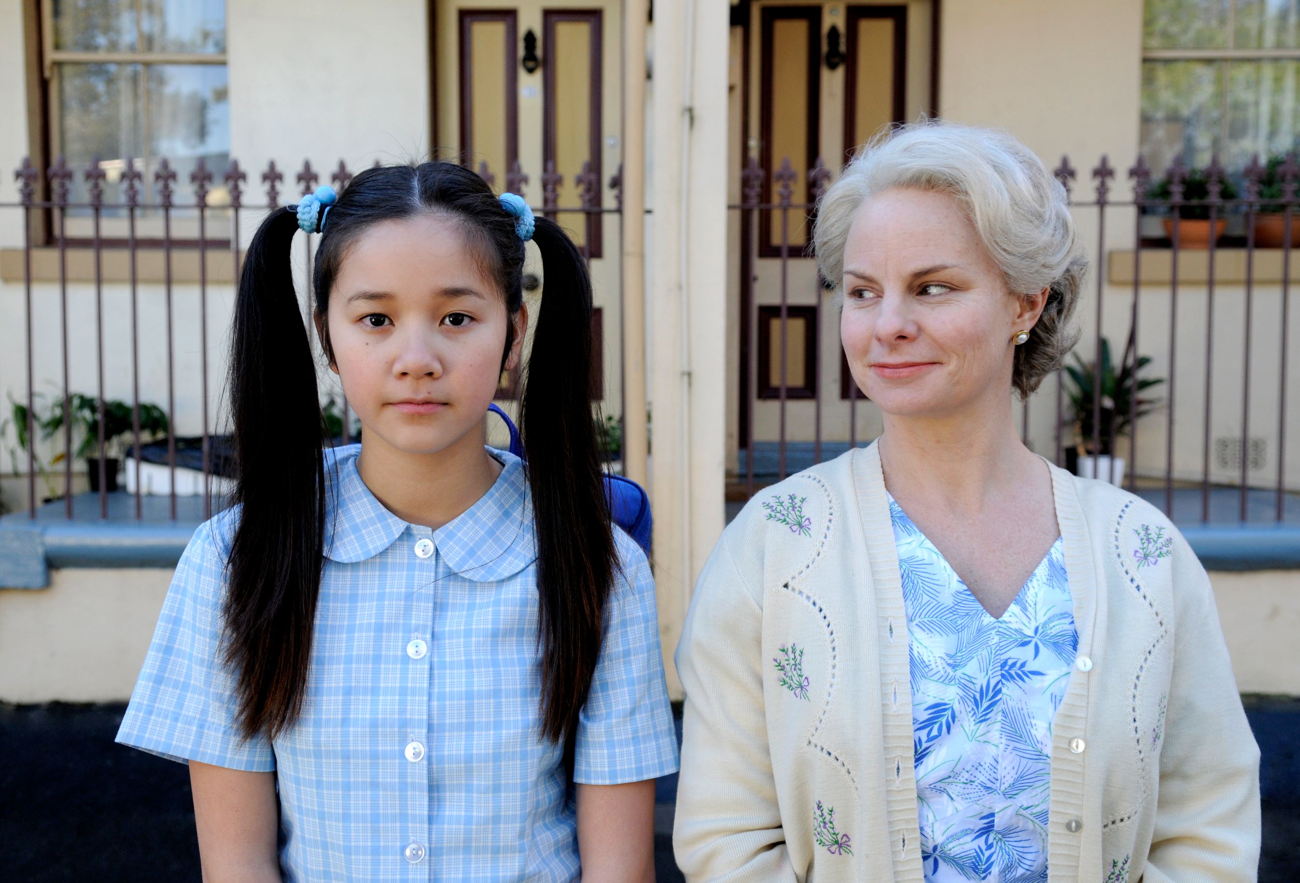 A young girl with long, dark hair tied into pigtails wears a blue checkered dress and looks solemn, standing next to an older woman with short, light hair and a warm smile, dressed in a floral blouse and cardigan. The background features a house with two doors and a decorative fence, hinting at a suburban setting.