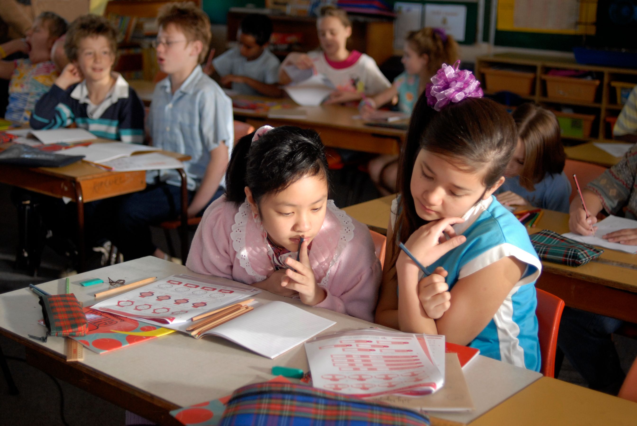 Two girls sit at a desk in a classroom, focused on their work. One girl wears a pink sweater with a lace collar and holds a pencil to her lips, while the other, dressed in a blue top, leans in to examine their papers, which feature various shapes and colors. In the background, several other students are engaged in their activities.