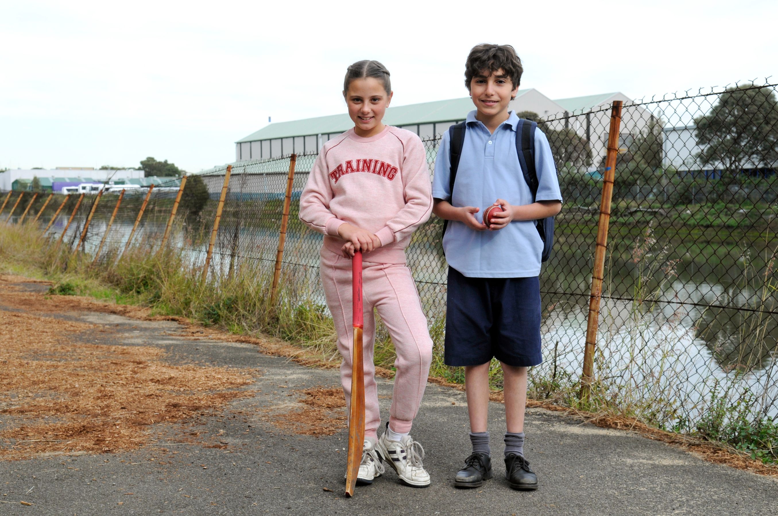 Two children stand on a path by a fenced area near a body of water. A girl in a pink tracksuit holds a cricket bat, while a boy in a blue shirt and shorts holds a cricket ball and has a backpack on.