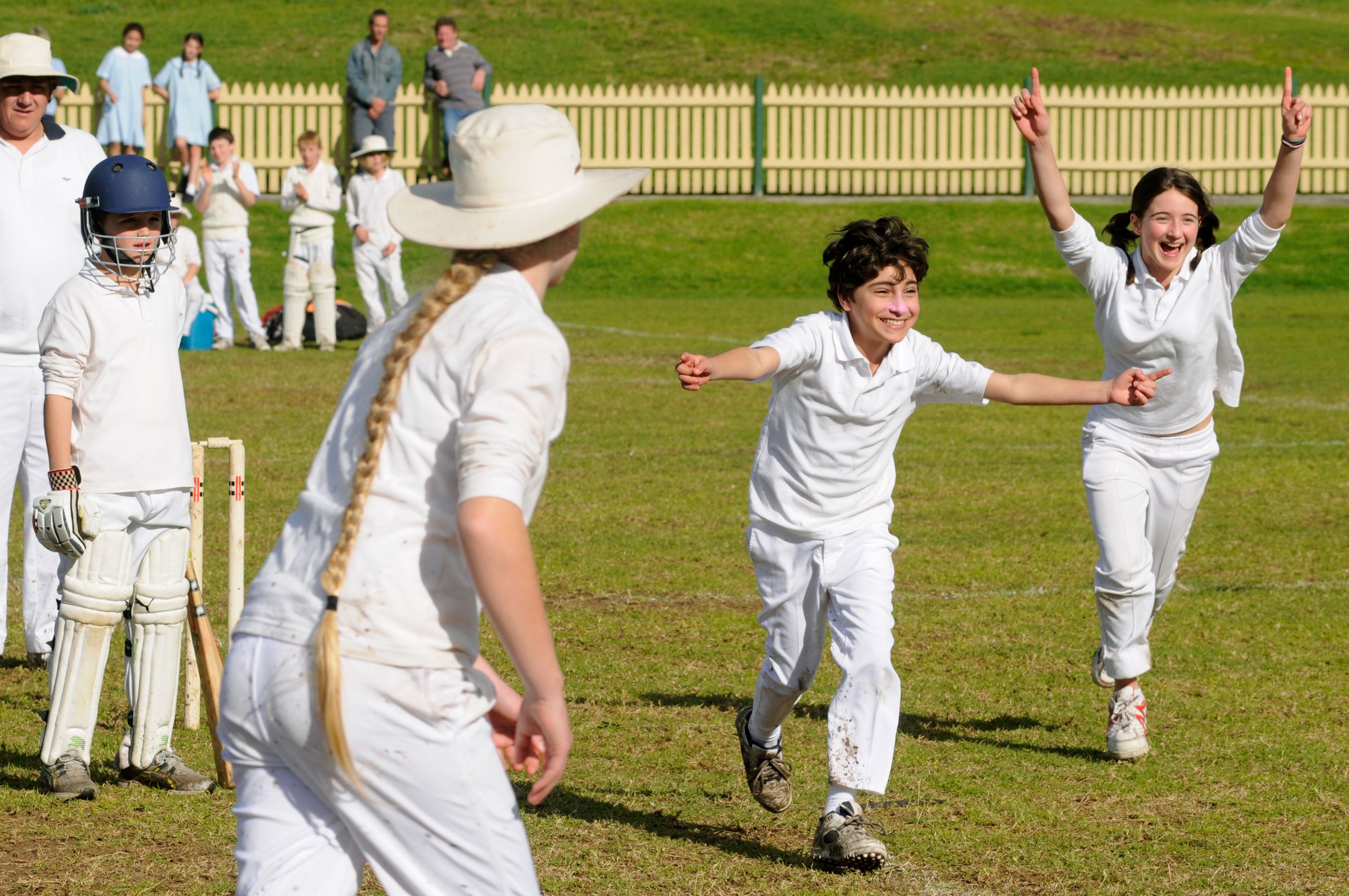 A group of children in white cricket uniforms celebrate joyfully in a grassy field, with two kids running towards the camera, arms raised in excitement. In the background, onlookers and players watch the celebration, highlighting a vibrant atmosphere of sport and camaraderie.