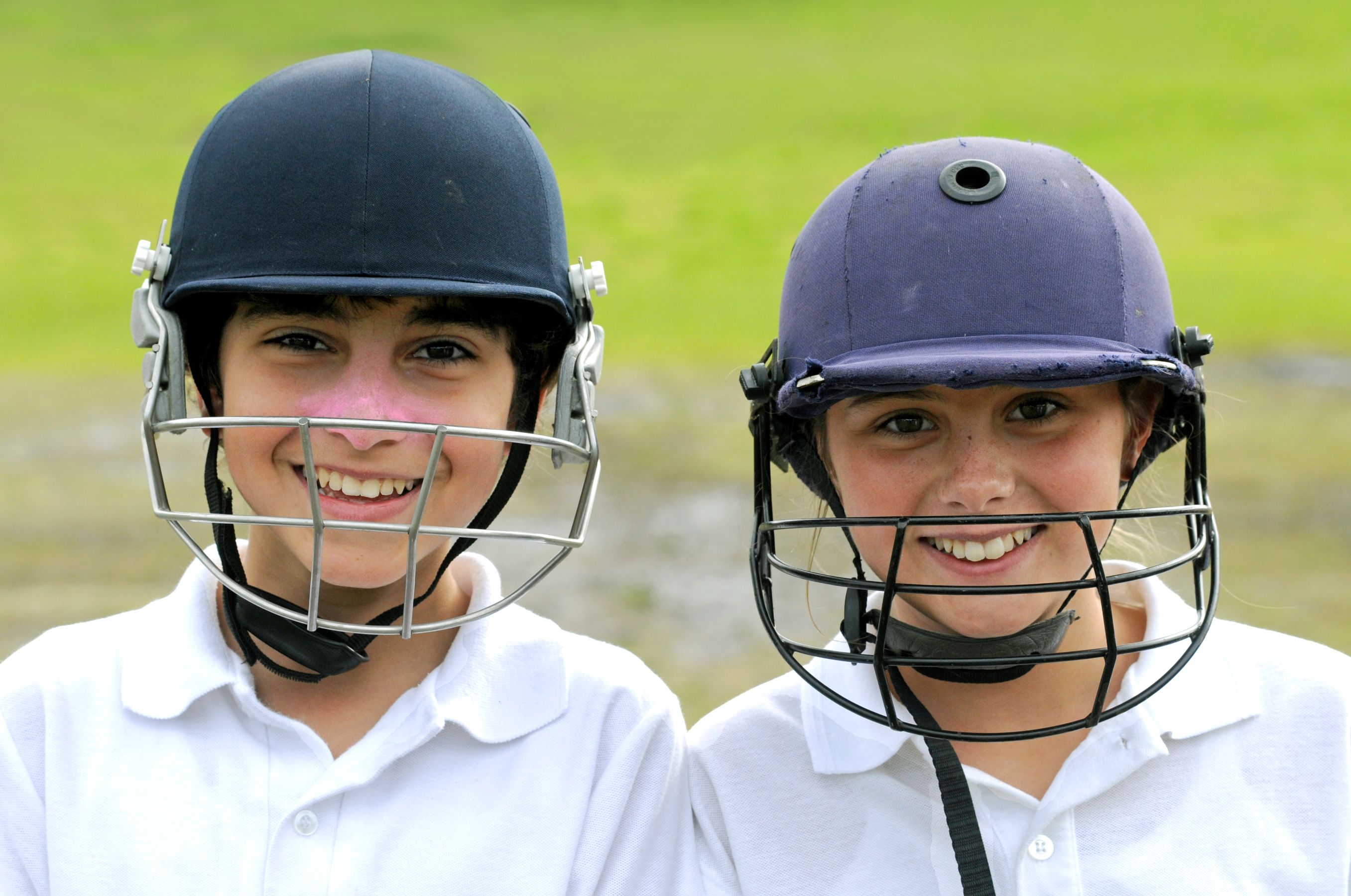 Two smiling young players wearing cricket helmets stand side by side, showcasing their enthusiasm for the sport. They are dressed in white shirts, and the background features a lush green field.