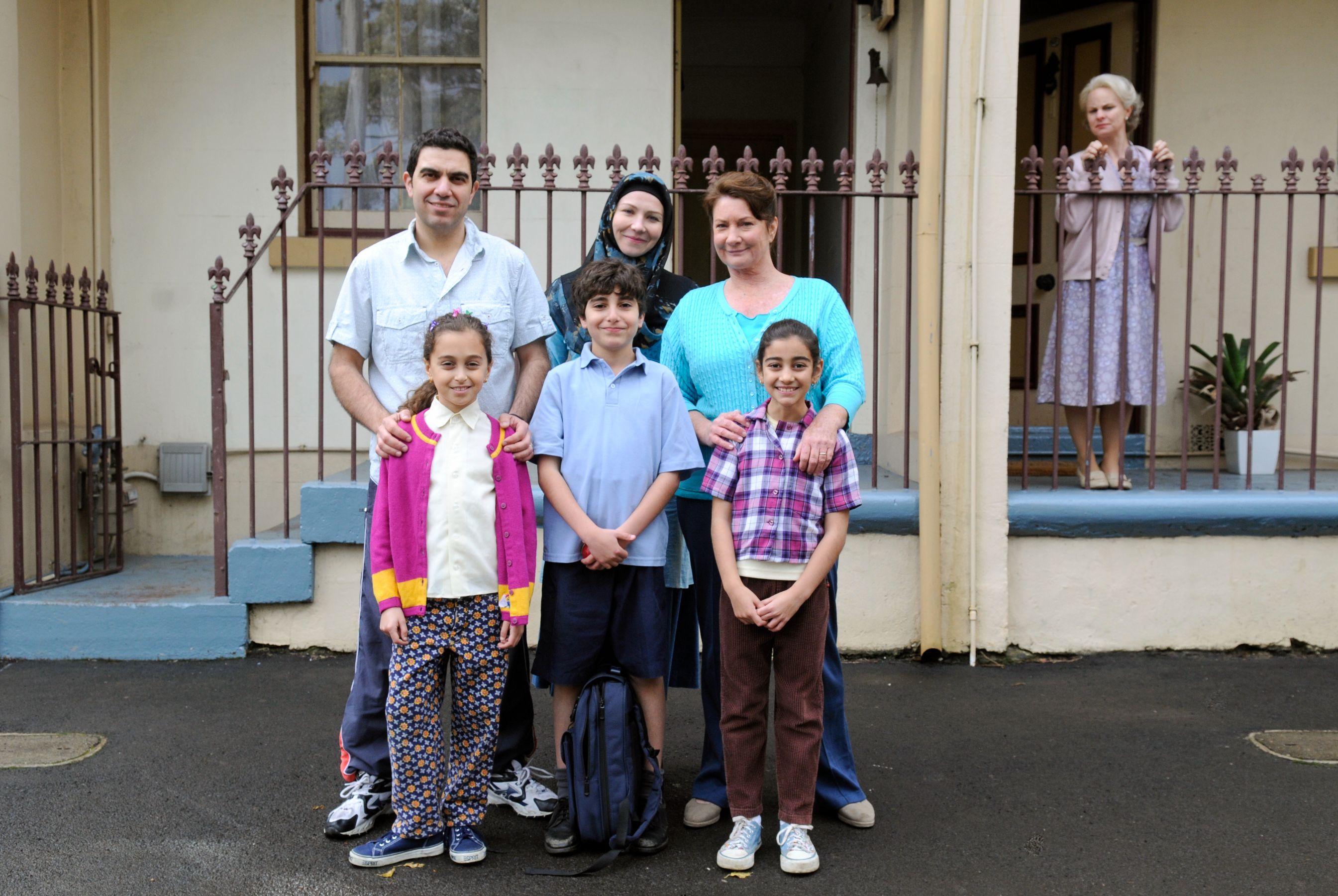 A diverse group of six people poses together in front of a house. The scene features three adults, two girls, and a boy, with the young children dressed in colorful clothing. The adults appear to be smiling and relaxed, while a woman stands in the background, looking on from the porch. The setting includes a well-kept yard and decorative plants.