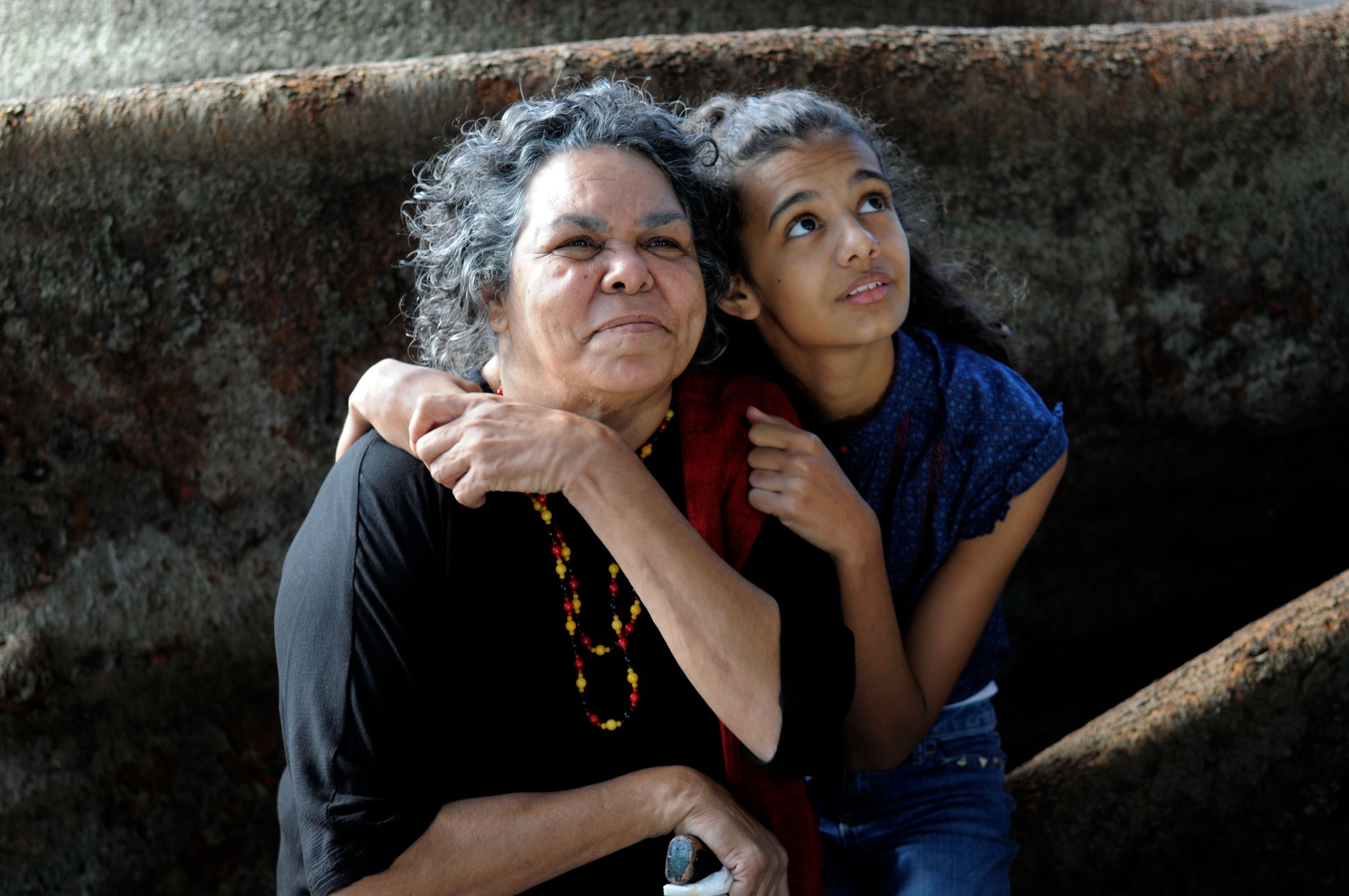 An elderly woman and a young girl share a moment of affection, with the girl resting her head on the woman's shoulder while looking upward with a thoughtful expression. The woman smiles gently, wearing a necklace of colorful beads, against a rustic backdrop.