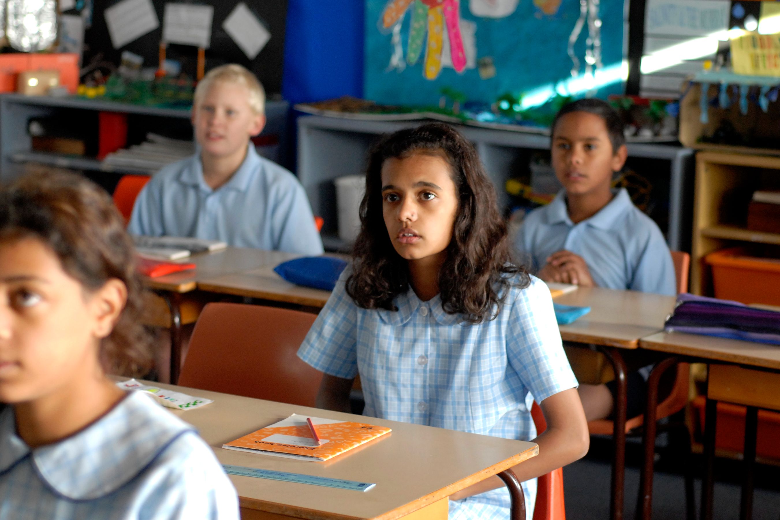 A classroom scene features several children seated at desks, wearing light blue school uniforms. The focus is on a girl with curly hair, looking attentively ahead, while other students are engaged in their seats. The background shows colorful educational materials on the walls.