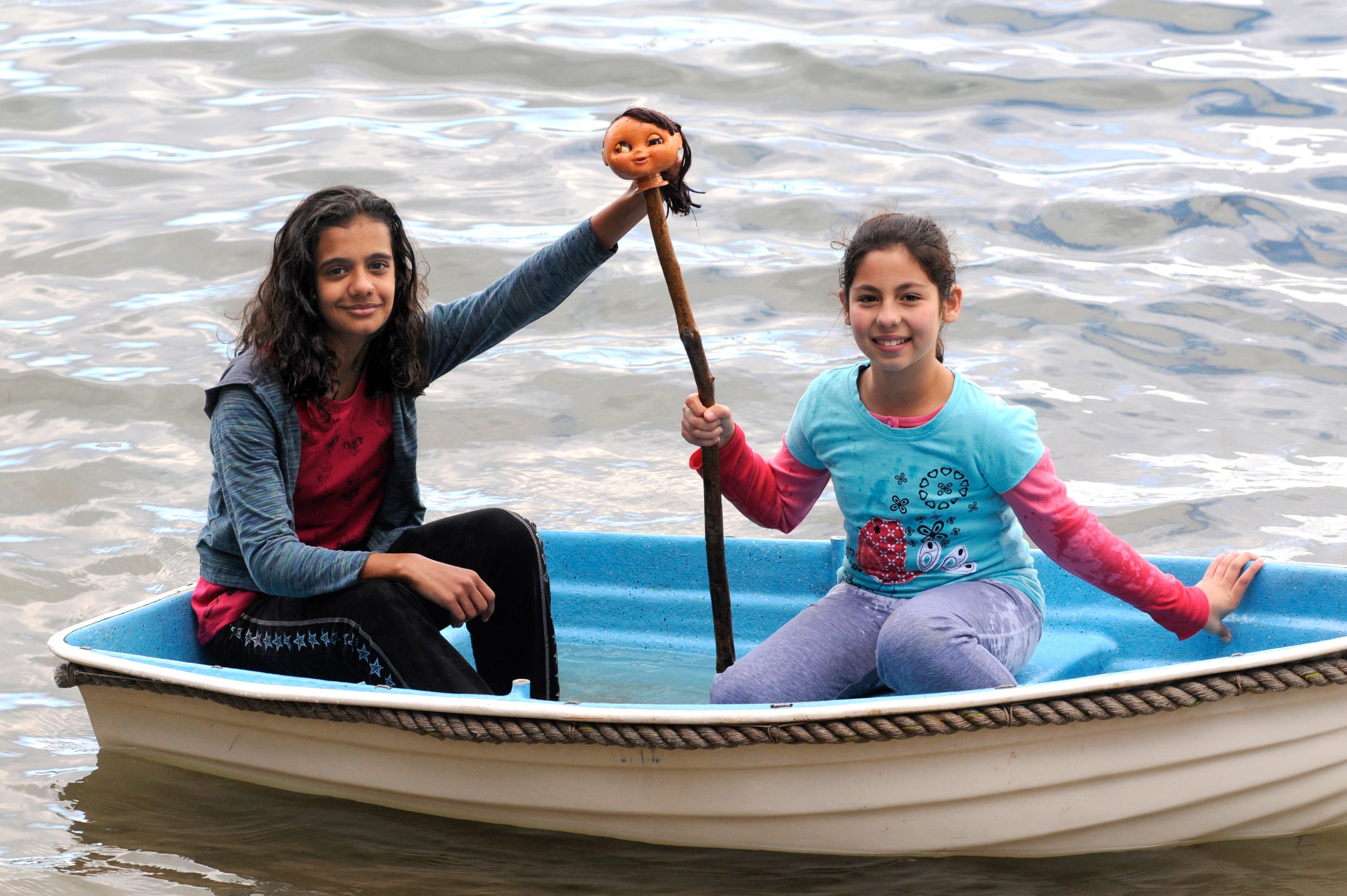 Two girls are sitting in a small boat on a body of water, smiling at the camera. One girl is holding a stick with a doll's head attached to it, while the other is sitting casually with her hand on the boat's edge. The water reflects the surroundings softly.