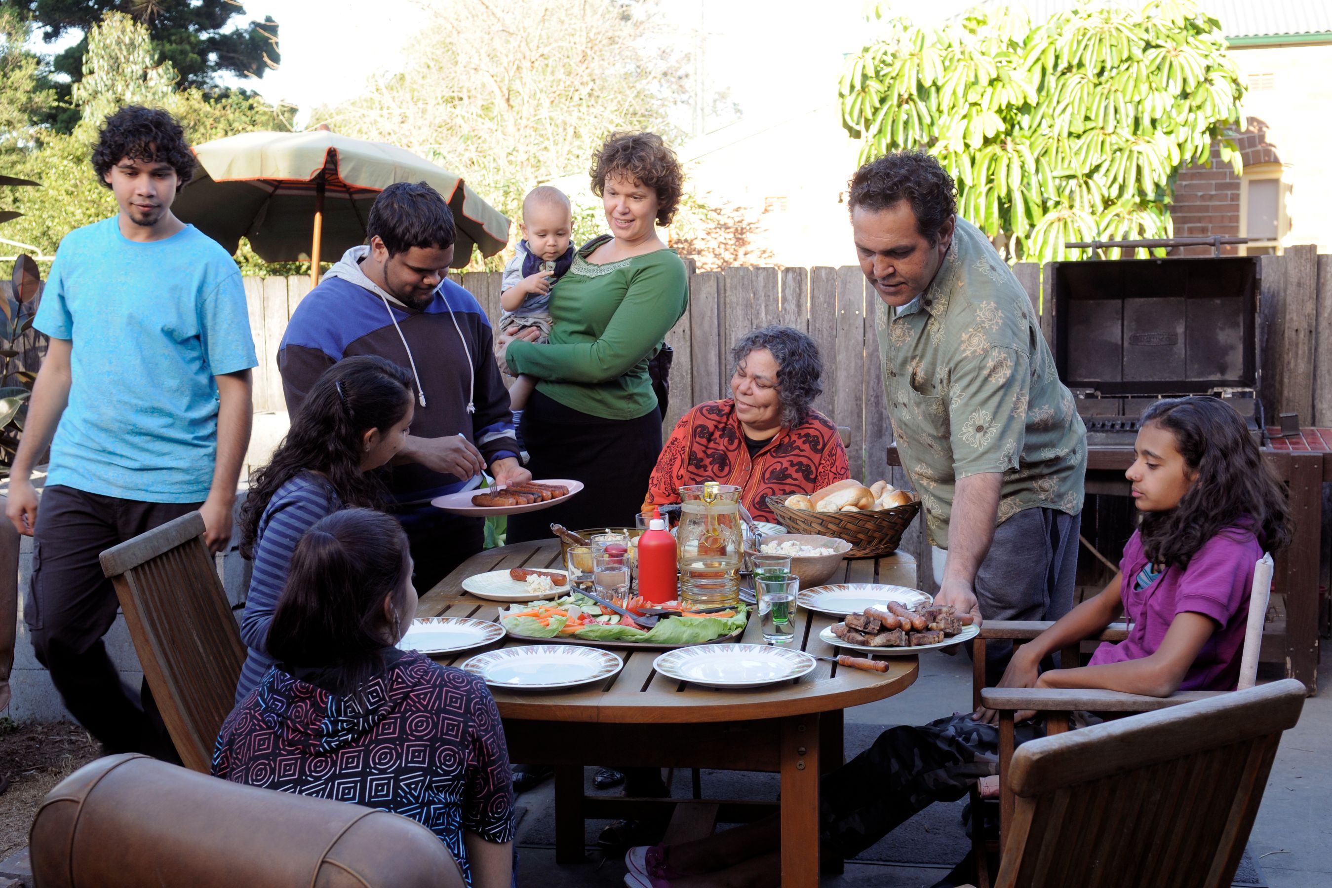 A diverse group of people gathers around a wooden table set with various dishes, including grilled meats and salads, in a backyard. Someone is serving food, while others chat and hold a baby, enjoying the outdoor setting surrounded by greenery and a grill in the background.