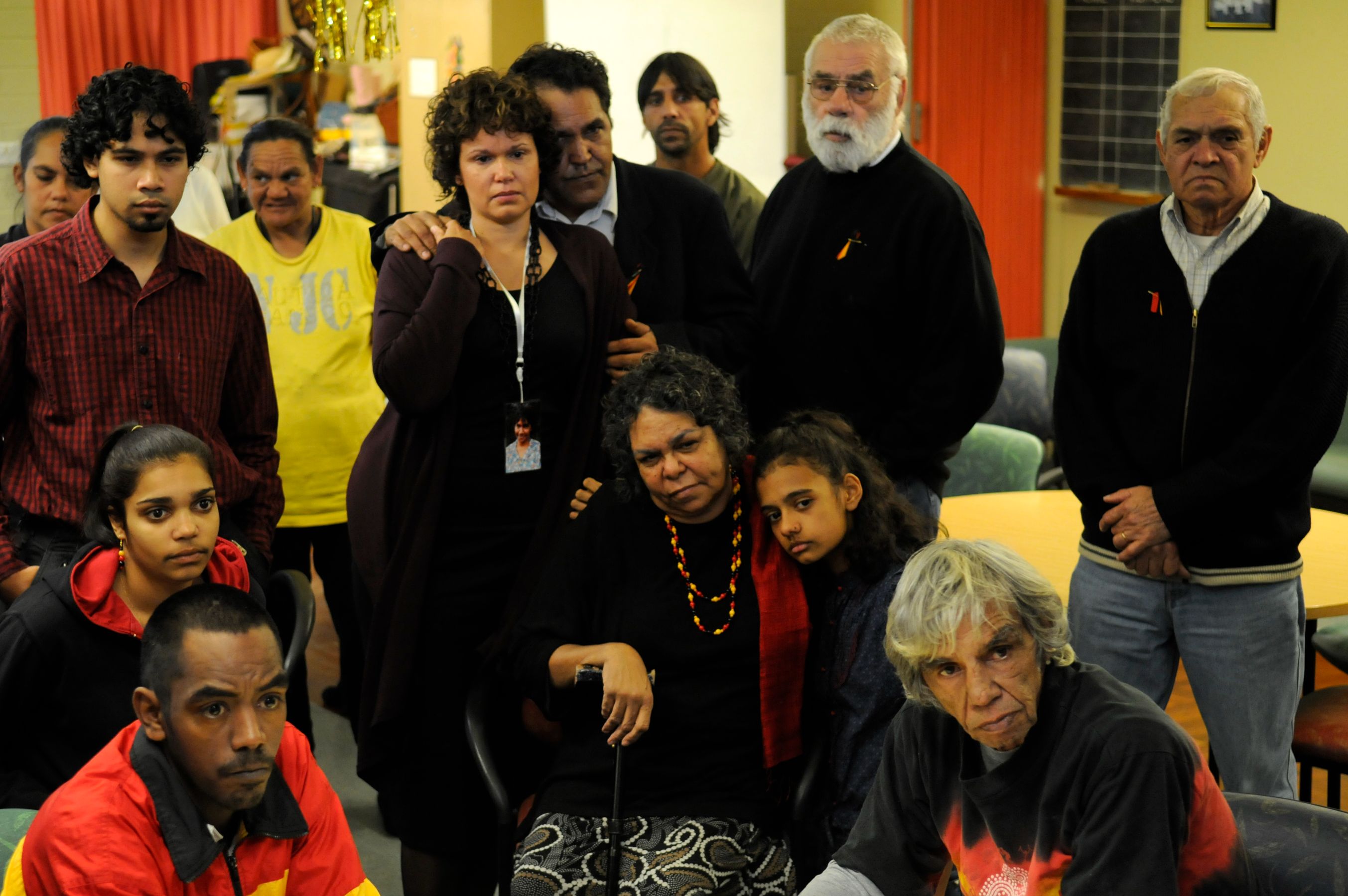 A diverse group of people stands and sits together in a communal space, showing expressions of concern and reflection. Some individuals are wearing colorful clothing, while others have more subdued attire, and there are items like a table and chairs in the background, suggesting a gathering or meeting environment.