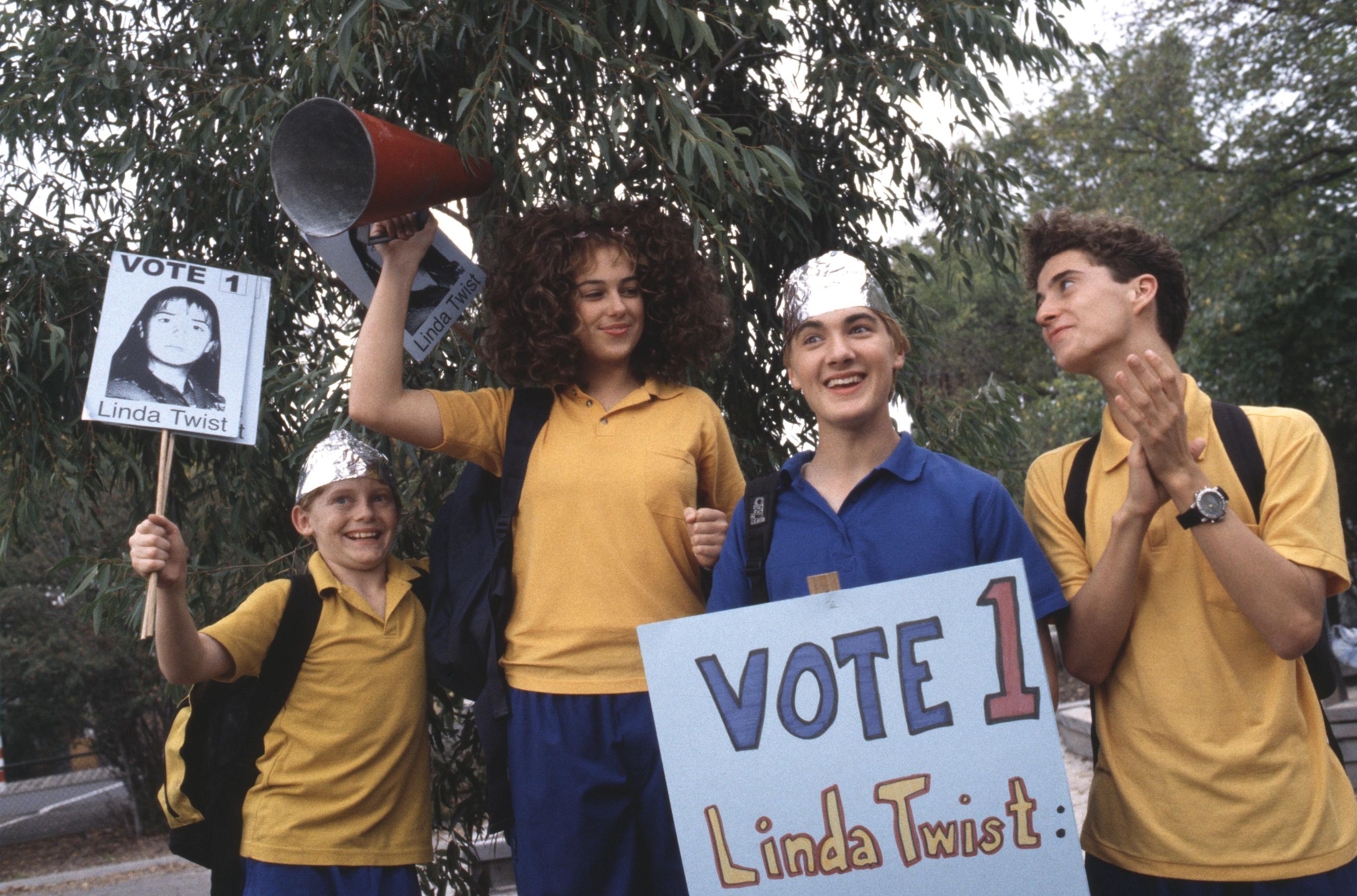 RTT_S4_EP07_2394 A group of four children stands outdoors, rallying for a campaign. One girl holds a megaphone high and wears a cheering expression, while another girl displays a sign with a photo of "Linda Twist." Two boys, one wearing a tinfoil hat, hold signs supporting the same candidate, smiling and looking enthusiastic. They are dressed in bright yellow and blue shirts.