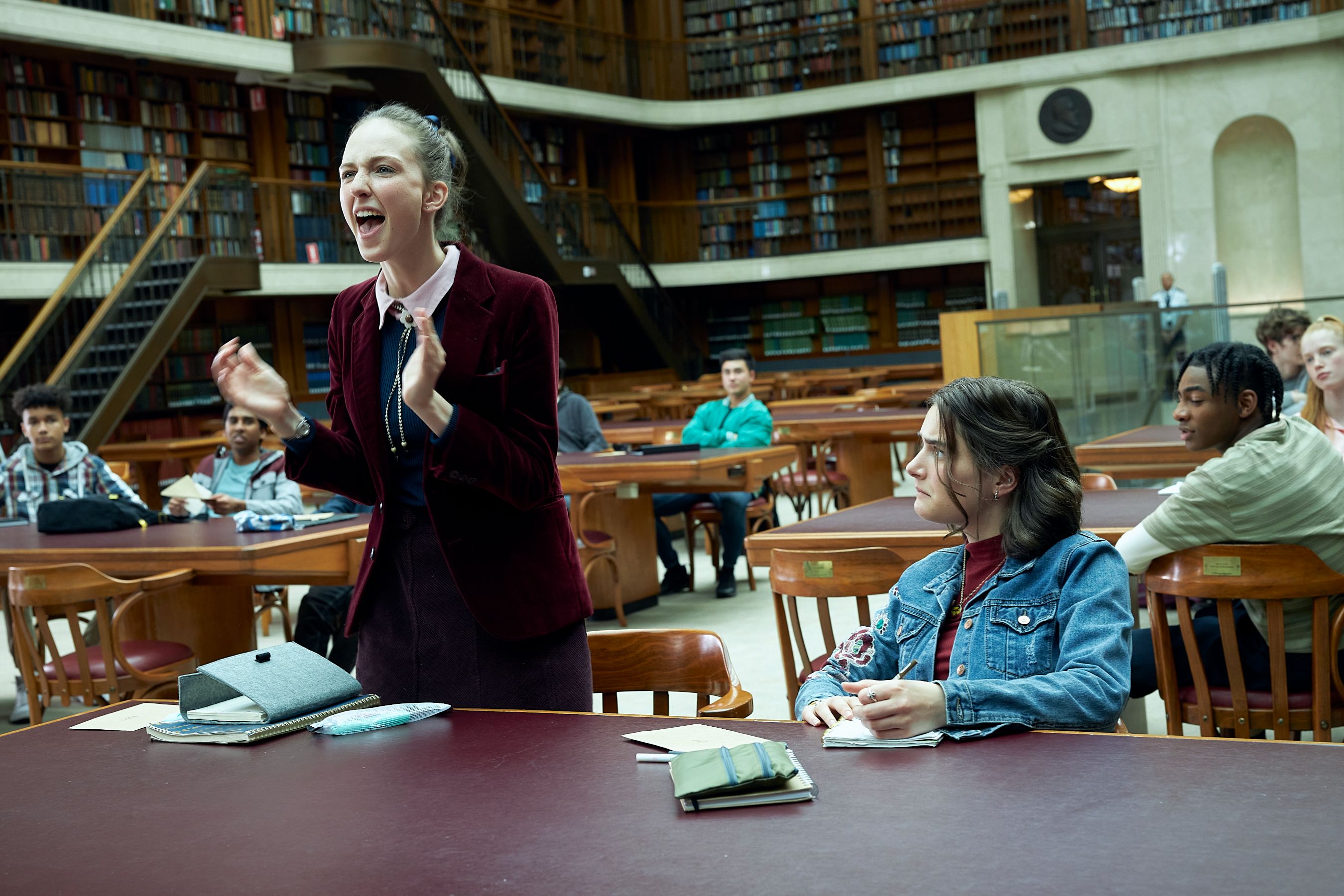 THE PM'S DAUGHTER_Sadie (Natalie English)_Cat (Cassandra Helmot)_Ep3_SD33_0105 A woman in a burgundy blazer passionately addresses a group of mostly young people seated at tables in a library, while they observe her attentively. The setting features wooden tables and shelves filled with books in the background.