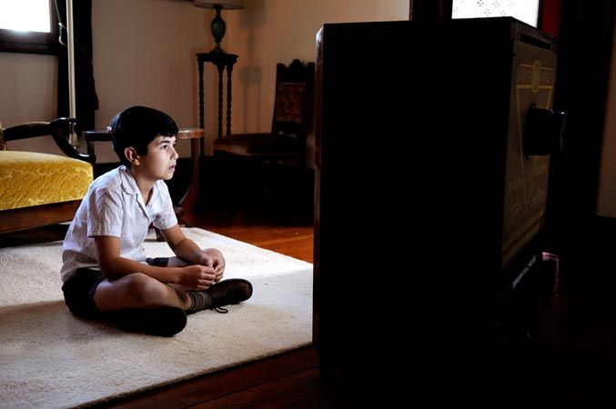 A young boy sits cross-legged on a rug in a dimly lit room, intently focused on an old television set in front of him. Behind him, a vintage chair and a table add to the nostalgic atmosphere of the space.
