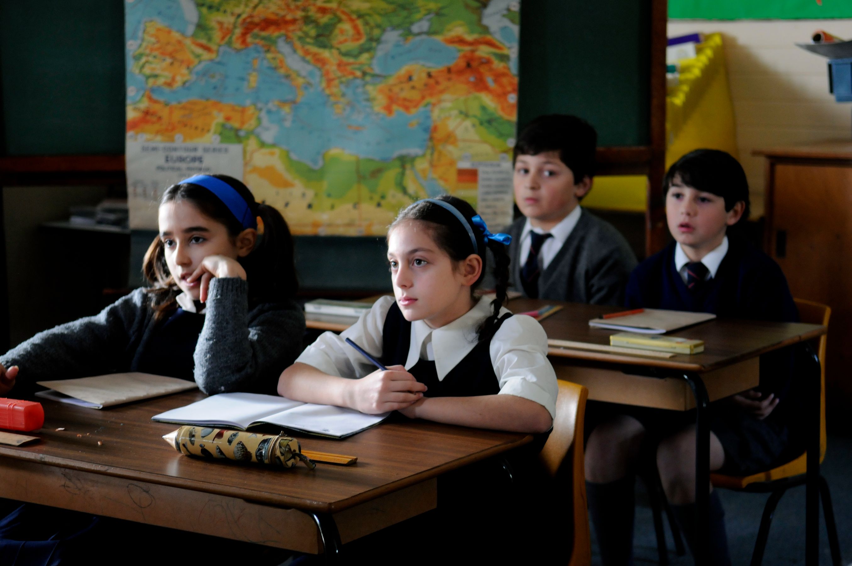 A group of four school children sits at wooden desks in a classroom, looking attentively toward the front. A large world map is displayed on the wall behind them, and their desks are scattered with notebooks and stationery. The children wear school uniforms, with two girls having their hair tied back with blue headbands.