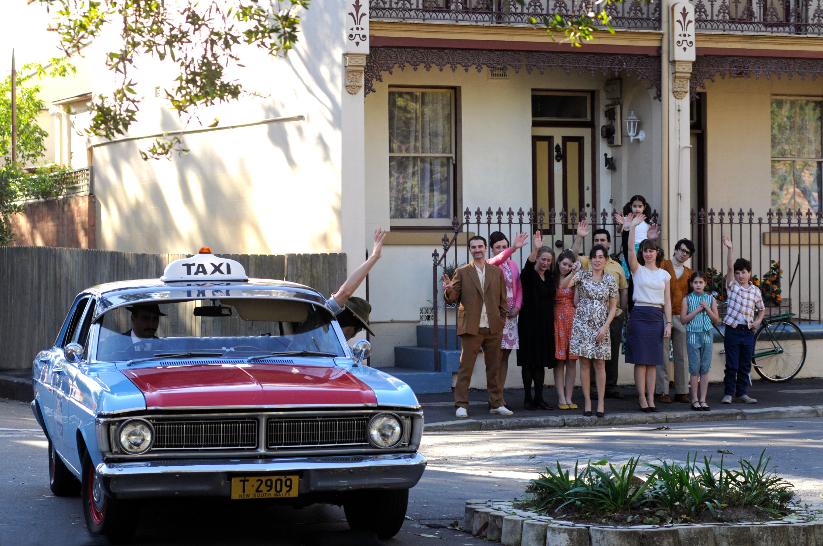 A blue and red taxi with a "TAXI" sign is parked on a street, while a diverse group of people, including men, women, and children, wave enthusiastically from the sidewalk in front of a two-story house. The scene captures a cheerful moment.