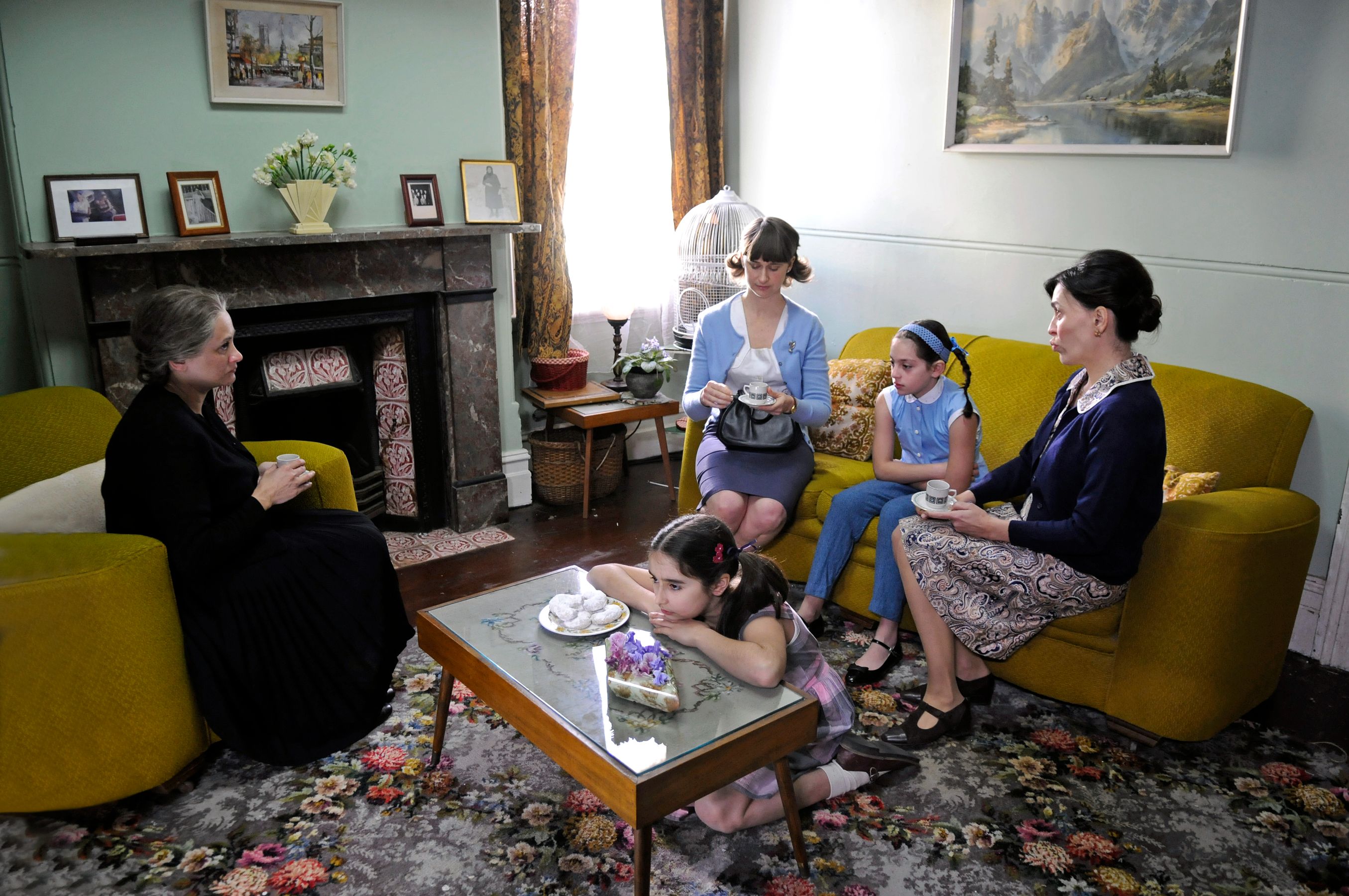 Five women of varying ages gather in a cozy living room, engaged in conversation over tea. The adults sit on a yellow couch while two young girls interact playfully on the floor, surrounded by patterned rugs and vintage decor, including a fireplace and photographs on the walls.