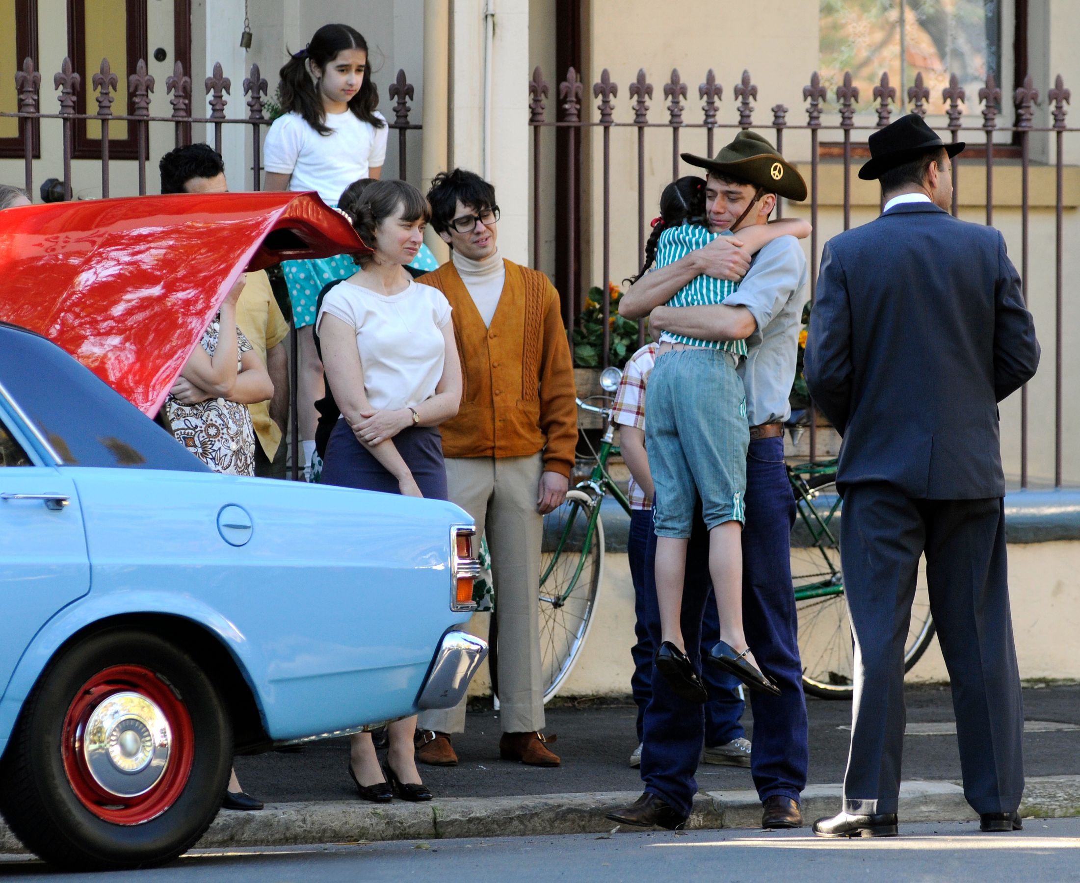 A group of people stands on a street, gathered around a blue car with its hood up. A man in a cowboy hat is holding a young girl, while others observe, including a woman with a relaxed expression and a boy wearing glasses. The background features a bicycle and a decorative fence.