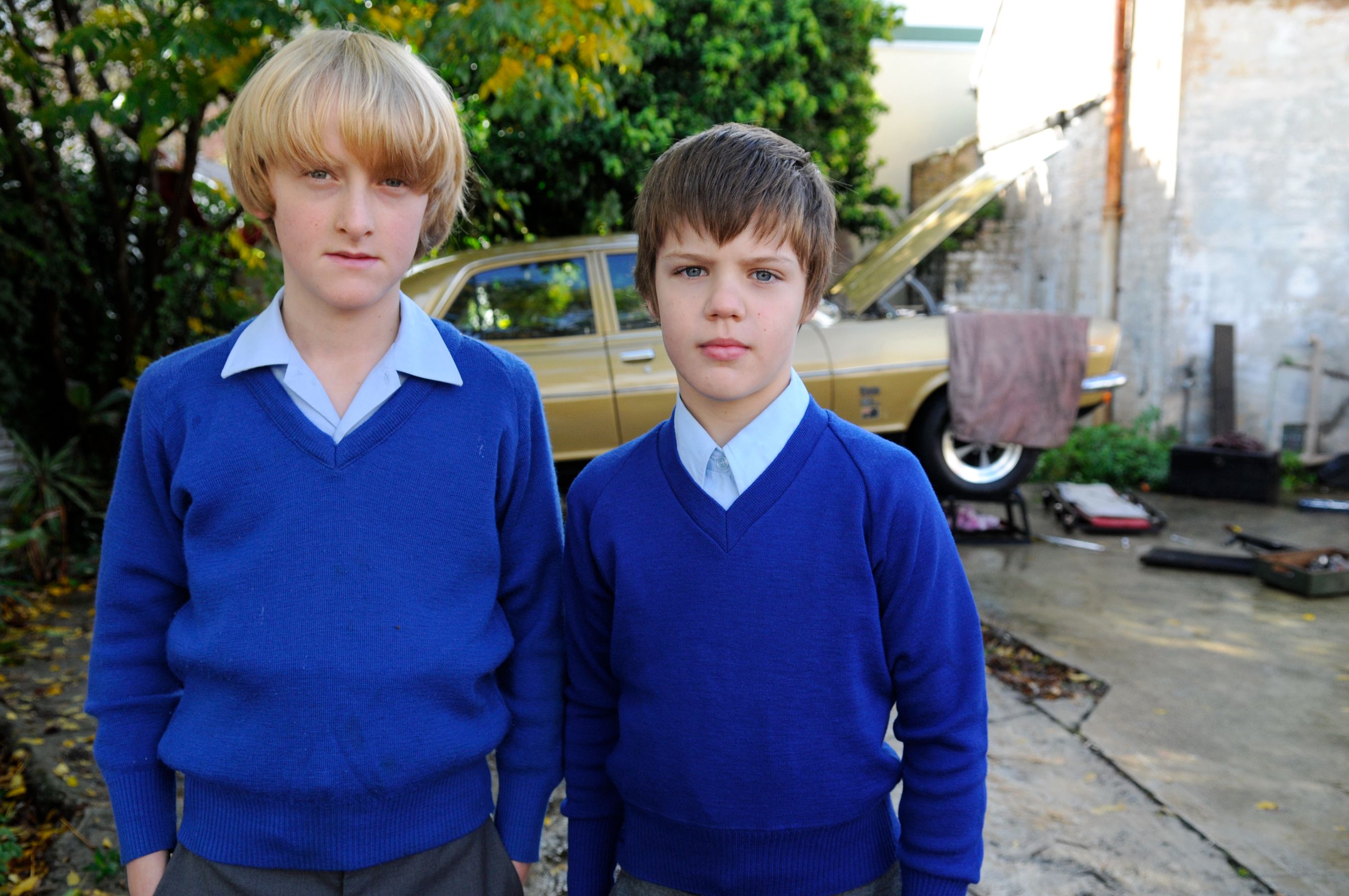 Two boys wearing blue sweaters stand together in an outdoor setting. Their serious expressions contrast with the vibrant greenery and a vintage car in the background, which has its hood raised. Various tools and parts are scattered on the ground nearby.