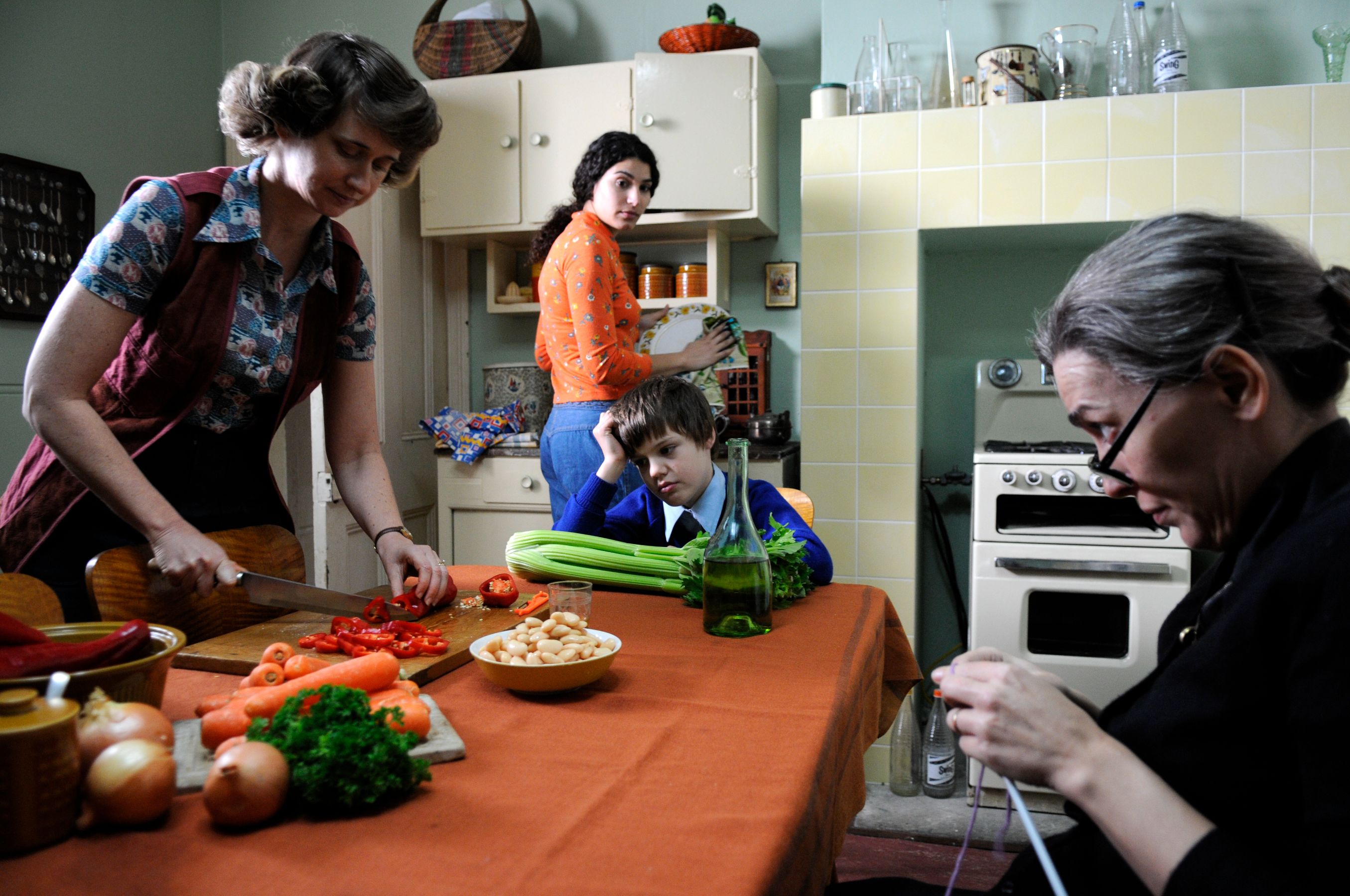A woman cuts vegetables at a table, while another woman in an orange sweater stands nearby holding a plate. A boy in a blue sweater looks frustrated, resting his head on his hand, and an older woman in black is seated, knitting. Various ingredients are laid out on the table, including vegetables and a bottle of olive oil, with a retro kitchen background.